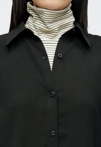 Crna košulja s ovratnikom s gumbima, slojevita preko prugaste rolke u krem i crnoj boji. Glatka tkanina s blagim sjajem.