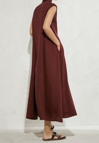 Ärmelloses Maxikleid in Burgunderrot mit Taschen, getragen von einer Person, die vor einer weißen strukturierten Wand steht, kombiniert mit schwarzen flachen Sandalen.