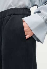 Main dans la poche d'un pantalon noir à taille élastique, portant un chemisier bleu clair à manches longues avec larges poignets, visible de la taille jusqu'au milieu de la cuisse.