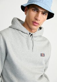 Felpa grigia con cappuccio e coulisse, dotata di un piccolo logo a patch. Tessuto morbido, caratterizzato da una vestibilità rilassata. Un cappello a secchiello blu chiaro completa l'outfit.