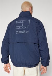 Marinblå vindjacka med elastiska ärmslut och nederkant, med en tryckt logotyp och textdesign på baksidan. Slät yta, lättviktsmaterial.