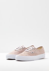 Un paio di sneakers Vans in suede rosa chiaro con lacci bianchi e spesse suole in gomma bianca su una superficie bianca.