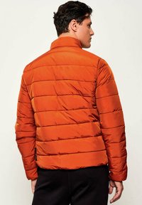 Giacca imbottita arancione con design trapuntato, colletto alto e polsini elasticizzati. Realizzata in materiale liscio e leggero con una vestibilità corta.