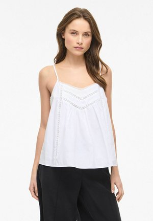 Młoda kobieta w białej bluzce bez rękawów z koronkowymi detalami i czarnych szerokich spodniach, stojąca na jednolitym białym tle.