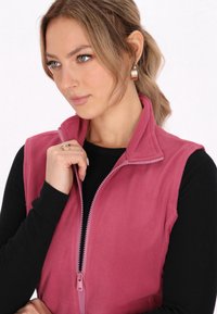 Gilet in felpa di un rosa chiaro, con colletto alto e cerniera frontale. Chi lo indossa tiene la cerniera mentre ha un maglione nero a maniche lunghe.