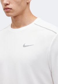 Κοντινό πλάνο ενός ατόμου που φοράει άσπρο μπλουζάκι Nike με γκρι λογότυπο swoosh και ανακλαστική λωρίδα στον ώμο.