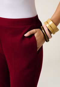 Bordeauxkleurige broeken met een gladde textuur, voorzien van zijzakken, gecombineerd met meerdere kleurrijke armbanden om één pols.