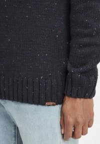 Marinblå stickad tröja med flerfärgade prickar, ribbade ärmslut och nederkant, med en liten läderetikett nära nederkanten.