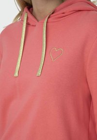 Sudadera color coral con cordones dorados y detalle de corazón bordado en el lado izquierdo del pecho. Material suave con un corte relajado.