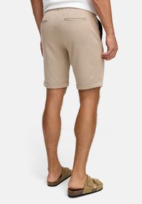 Shorts beiges en tissu texturé, avec des revers roulés et deux poches arrière. Portés avec des sandales marron clair à lanières croisées.