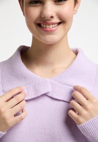 Maglione lilac lavorato a maglia con colletto a costine e un corpo testurizzato. Le mani del modello sistemano il colletto, mostrando il tessuto morbido.