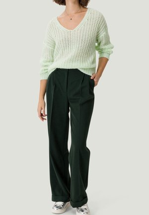 Femme portant un pull léger en tricot vert clair, un pantalon large vert foncé et des baskets métalliques argentées, debout avec une main dans la poche.