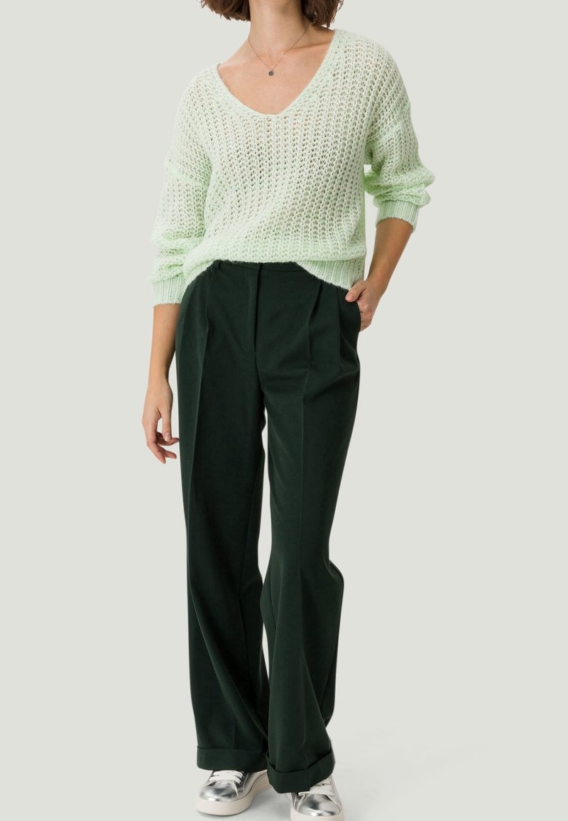 Femme portant un pull léger en tricot vert clair, un pantalon large vert foncé et des baskets métalliques argentées, debout avec une main dans la poche.