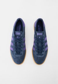 Sötétkék velúr sportcipők lila csíkkal és gumitalppal. Fűzős kivitel és párnázott szegély a kényelem érdekében.