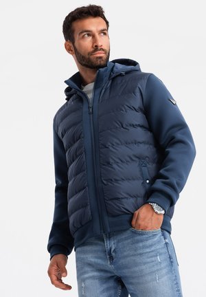 Chaqueta de pluma navy con capucha, que presenta un diseño acolchado, cierre frontal con cremallera y puños elásticos. Combinada con jeans desgastados de color azul claro.