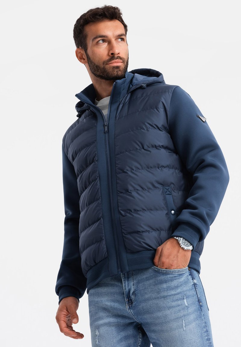 Marinfärgad puffjacka med huva, med quiltat mönster, dragkedja fram och elastiska manschetter. Kombinerad med ljusblå slitna jeans.