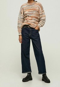 Personne portant un pull en maille à rayures multicolores, un jean large noir avec une ceinture brune et des bottes noires épaisses sur fond uni.