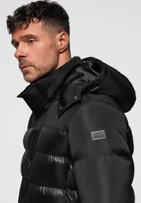 Giacca a vento nera con collo alto, cappuccio regolabile e finitura lucida. Patch del logo ben visibile sulla manica sinistra, design a texture trapuntata.