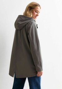 Chaqueta gris, impermeable, con capucha, de corte relajado y aberturas laterales. Textura suave y hardware mínimo. Combinada con denim azul.