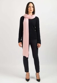 Una sciarpa rosa chiaro avvolge il collo; materiale in lana con una texture morbida. Abbinata a un maglione nero aderente e pantaloni skinny neri.
