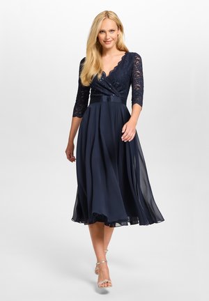 Femme portant une robe bleu marine en dentelle avec manches trois-quarts et une jupe fluide, marchant vers l'avant sur un fond clair uni.
