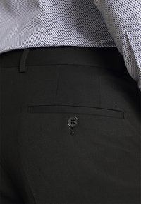 Pantaloni neri su misura caratterizzati da una texture liscia, una tasca posteriore con un bottone nero e una leggera lucentezza. La cintura è piatta e dotata di passanti per cintura.