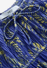Tissu à motifs bleu et jaune avec une finition texturée. Présente une taille nouée et une étiquette indiquant "NEXT". Design doux et froncé.