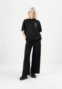 T-shirt nera oversize con stampa bianca, abbinata a pantaloni neri larghi. Il modello indossa sneakers nere. Design minimalista su sfondo bianco.