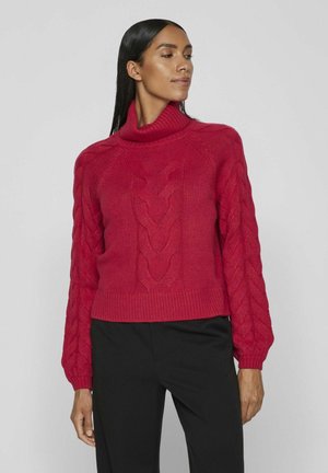 Rød strikket sweater med høj ribbet krave, kabelstrik-design på ærmerne og kort længde. Båret med sorte bukser.