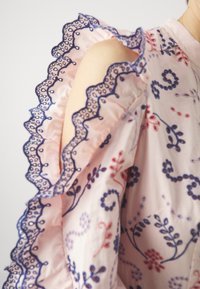 Gewebte Bluse aus weichem, pinkfarbenem Stoff, mit floralem Stickmuster in Blau und Rot. Rüschensleeves mit gewelltem blauen Saum betonen das Design.