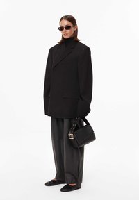 Czarna, oversize marynarka z subtelnymi detalami kieszeni, zestawiona z szerokimi, czarnymi skórzanymi spodniami, płaskimi czarnymi butami i usztywnioną czarną torebką.