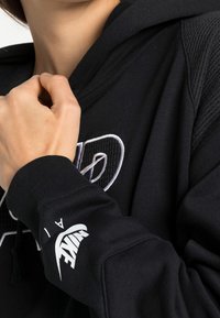 Person med en svart Nike-huvtröja, hand som justerar kragen, visar Nike-logotypen och broderad design på bröstet och ärmen.