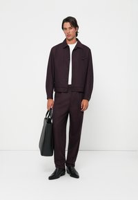 Burgundy textured jacket with black buttons, paired with matching trousers and a white t-shirt; model holds a black tote bag.