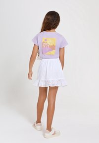 Fialové tričko s grafickým potiskem palmy na zadní straně, v kombinaci s bílou sukní z očkované látky s volánkovým lemem. Oblečeno s lehkými teniskami.