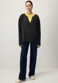 Pull gris foncé à col en V avec un col jaune, orné de trois boutons marron. Associé à un jean bleu ample et des chaussures de couleur claire.