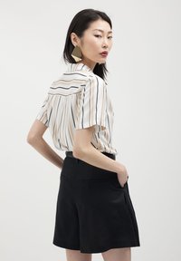 Donna con lunghi capelli scuri, che indossa grandi orecchini geometrici dorati, una camicia bianca maniche corte con strisce beige e nere e pantaloni corti neri a vita alta.