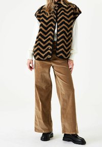 Faux päls väst med chevronmönster i svart och tan, kombinerad med bruna vida kordbyxor och svarta plattformsskor.