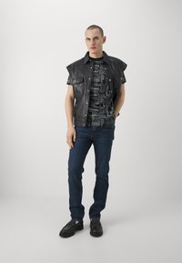 Chaleco de cuero negro sin mangas sobre una camiseta gráfica negra, jeans azul oscuro y mocasines negros. Diseño simple y moderno con materiales contrastantes.