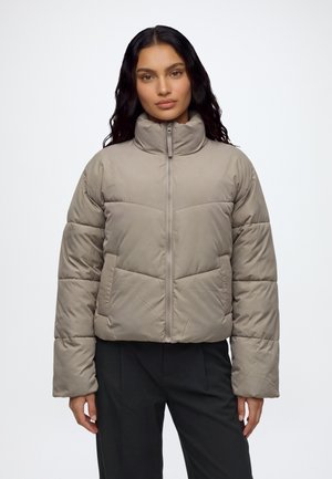 Femme aux longs cheveux foncés portant une veste matelassée gris clair avec un col montant et un pantalon noir sur fond blanc uni.