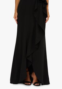 Czarna długa sukienka z falbaną po boku, wykonana z gładkiego materiału, z odsłoniętymi palcami w czarnych sandałach na paski.
