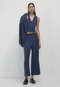 Marineblaues maßgeschneidertes Anzugset mit einem kurzen Blazer mit drei Knöpfen und weitem Hosenbein. Das Modell hält einen passenden Blazer. Glänzende schwarze Mokassins.