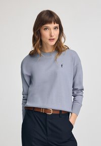 Pull léger gris en matériau doux avec un col rond et des poignets côtelés, présentant un petit logo marine sur la poitrine. Accent ceinture marron.