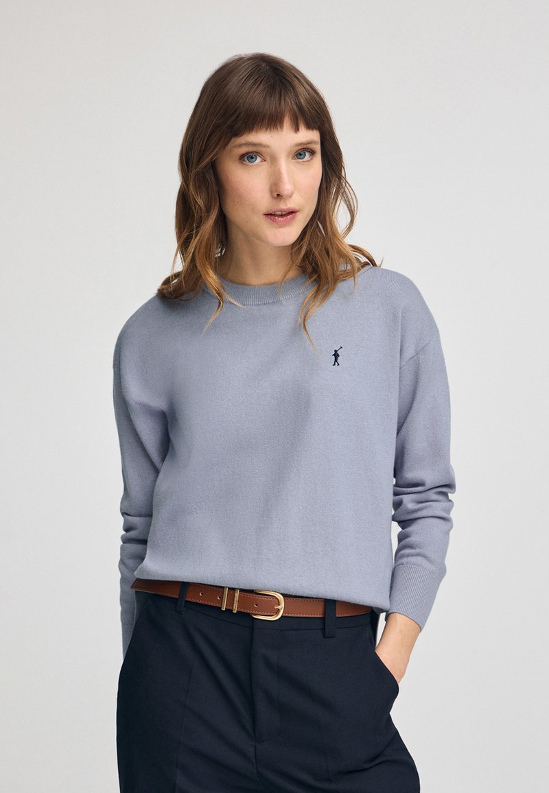 Pull léger gris en matériau doux avec un col rond et des poignets côtelés, présentant un petit logo marine sur la poitrine. Accent ceinture marron.