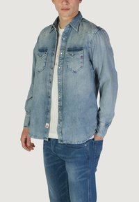 Camicia di denim azzurro chiaro con bottoni a pressione, due tasche sul petto e un yoke in stile western. Indossata sopra una maglietta bianca, abbinata a jeans blu.