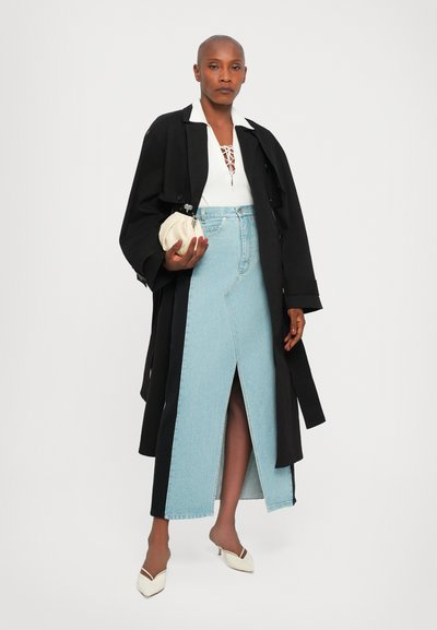 Cappotto trench nero sopra una maglietta bianca con dettagli in pizzo, abbinato a una gonna di jeans azzurri chiari con uno spacco alto fino alla coscia e tacchi beige.