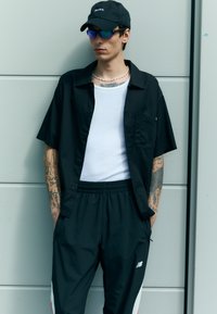 Black short-sleeve button-up shirt over a white tank top, paired with black elastic-waisted pants featuring red and white stripes, sunglasses, and a cap.