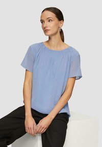 Helles blaues kurzärmeliges Blusenshirt mit rundem Halsausschnitt und gerafften Details. Leichtes, fließendes Material; kombiniert mit schwarzen Hosen.