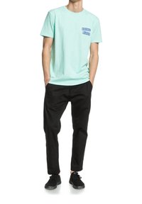 Muž oblečený v světle modrém tričku "Vacant Beach", černých kalhotách a černých teniskách, stojící s rukama v kapsách na bílém pozadí.