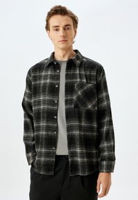 Camicia in flanella a quadri bianca e nera con bottone frontale, maniche lunghe, taschino singolo sul petto e strato interno grigio. Tessuto morbido.