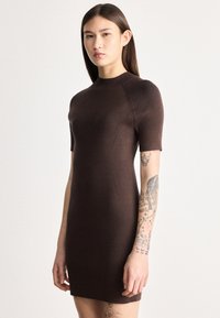 Γυναίκα με μακριά σκούρα μαλλιά, ντυμένη με εφαρμοστό καφέ ριμπ μπλουζάκι με κοντά μανίκια, που δείχνει λεπτομ�ερείς τατουάζ στο δεξί της χέρι και πόδι.